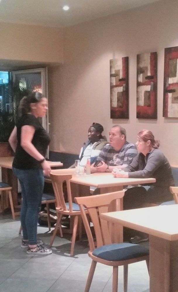 A standing woman addresses three people, two women and a man, all seated, oddly, in a row. They all have expressions of hopelessness on their faces. 