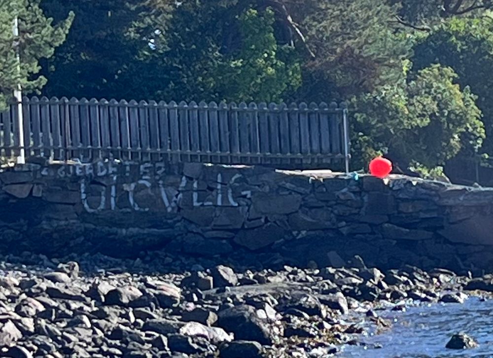 Et bilde av en steinstrand med en steinmur i bakgrunnen med et gjerde på seg. Dette gjør strandsonen mer utilgjengelig for folk, og på steinmuren er det tagget "gjerdet er ulovlig"
