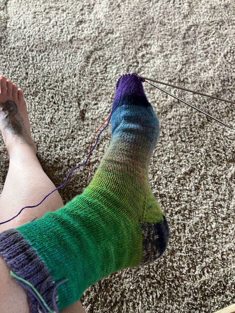 Very colorful hand knit sock that I was trying on which was almost finished knit. Greens, yellow, blue, purple, dark grey in color. 