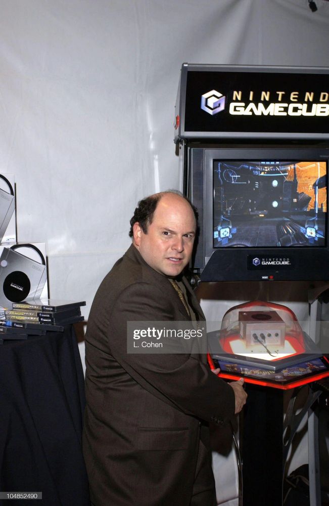 Jason Alexander looking to camera like a cornered animal as he plays a display model Nintendo GameCube.