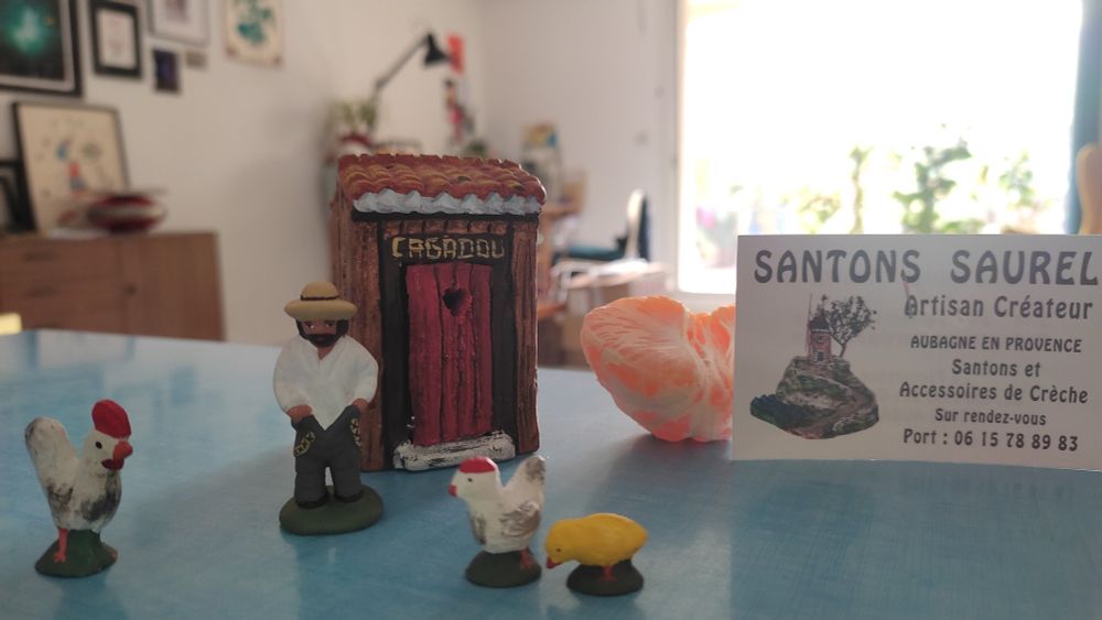 Un santon (figurine en terre cuite typique en Provence) qui sort du cagadou (petite cabane pour faire ses besoins, en parlé marseillais), un poussin, une poule et un coq. 
Une demie clémentine sur laquelle s'appuie la carte de l'artisan : Santon Saurel 