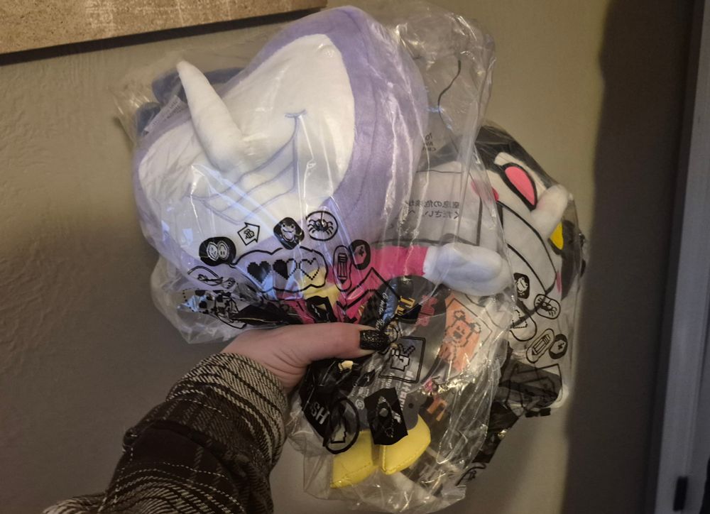 Picture of me holding a Tenna plush and a Spamton plush in one hand. Both are still in the plastic bags they were shipped in.