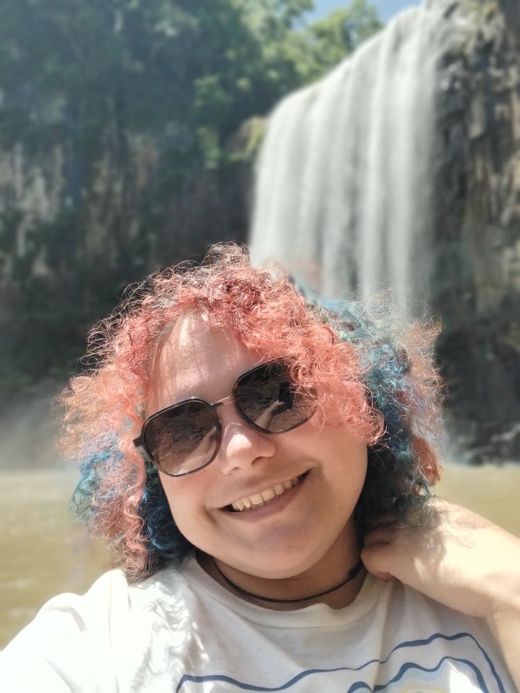 Mulher de óculos escuro, cabelo cacheado vermelho com mechas azuis, blusa branca, sorrindo em frente a uma cascata em um dia ensolarado 