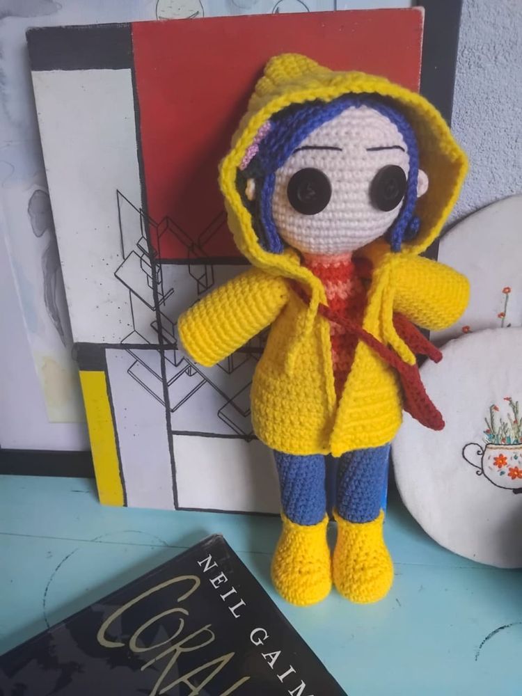 Boneca de crochê da Coraline, menina branca de cabelo azul, olhos de botão pretos, casaco amarelo com capuz, calça azul, bota amarela, bolsa vermelha. Ao fundo quadro vermelho, cinza e azul, bordado de um açucareiro com plantas dentro. No chão o livro da Coraline.