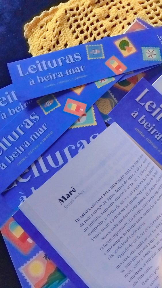 Livros azuis espalhados sobre mesa preta com trilho amarelo. O livro aberto em primeiro plano tem o conto Maré, de Jozieli Weber, aparecendo. Marcadores de página com o título do livro: Leituras à beira mar, e desenhos de selos postais, estão espalhados sobre os livros.