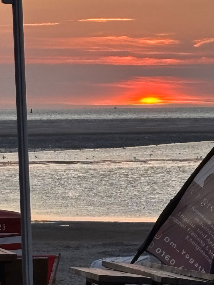 Sonnenuntergang im Meer, davor eine Sandbank. Am linken und rechten Rand eine Stange und ein Segelbanner. 