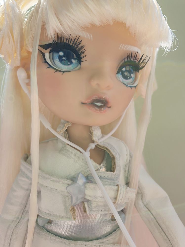 photo of the Rainbow High doll Whitney, who has long platinum blonde hair with two small twintails, black sparkly lipstick, a white pleather jacket and skirt over a metallic silver bodysuit, and wired headphones attached to a retro portable cd player. the light is washed out because my camera phone didn't like how bright the sunlight was today.