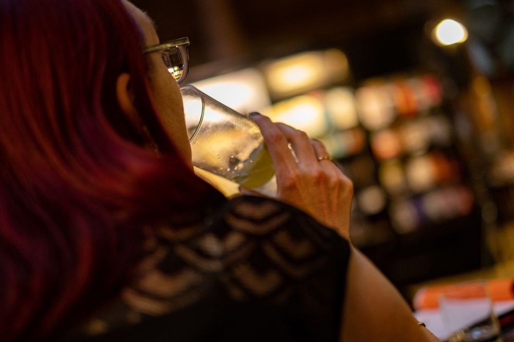 We look over the shoulder of a purple haired lady who is drinking a cocktail. Jazz plays in the background 