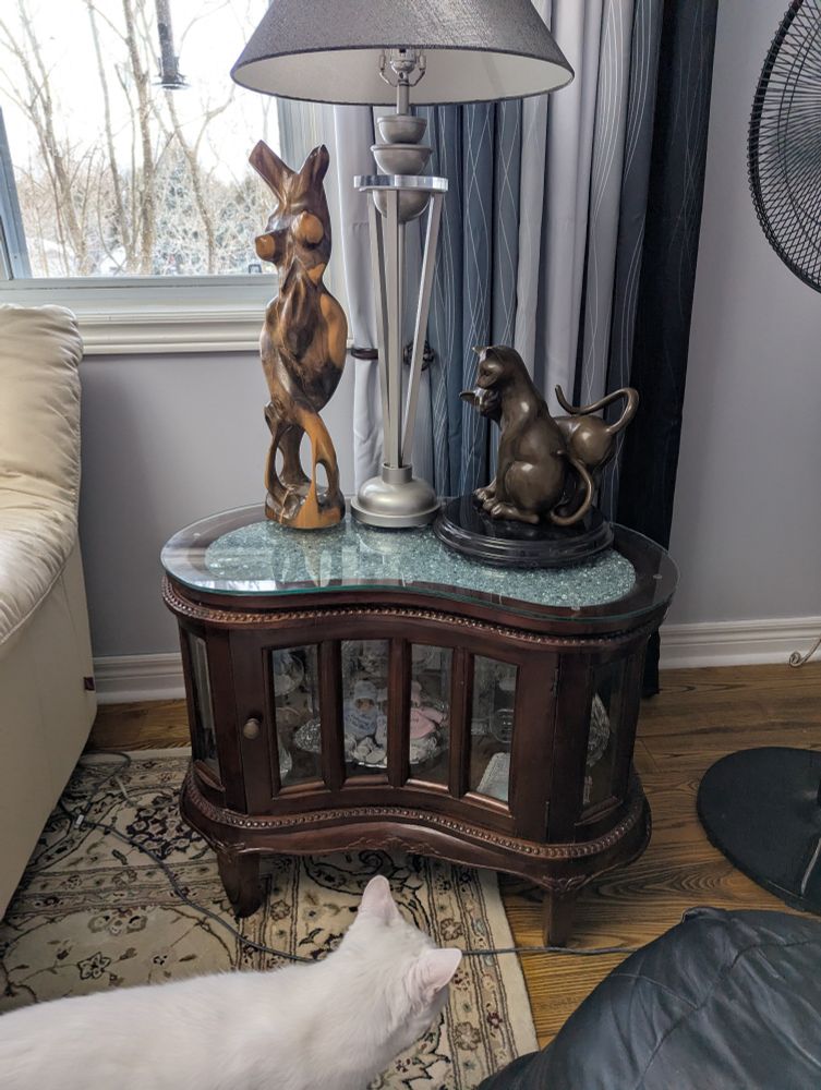 The cat is very interested in the new addition to the living room - especially the small items behind the glass cabinet doors. Items on top of the table are: a carved wood abstract figure of a woman, stainless steel and lucite lamp from the early 1980s, and a hot cast bronze sculpture of two intertwined cats on a black marble base.
Partial of a white couch side, black ottoman, window, and curtains also in image.