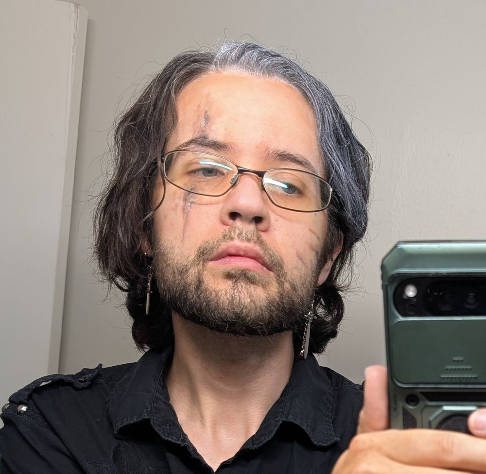 A mirror selfie of a man in Verso (Expedition 33) costume makeup with white spray-dye in the relevant place. He is wearing sword earrings and a black button-up shirt.