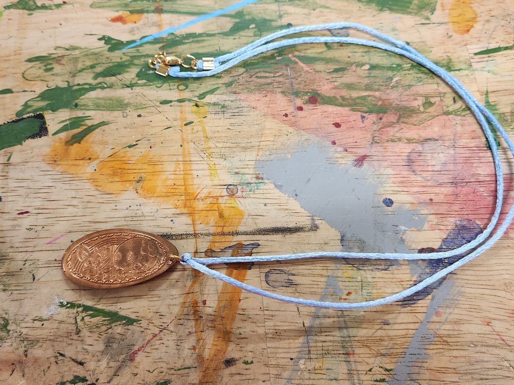 The pressed coin from the previous image, but now on a light blue cord necklace as a pendant! The clasps and jump rings are a matching metal color to the coin. 