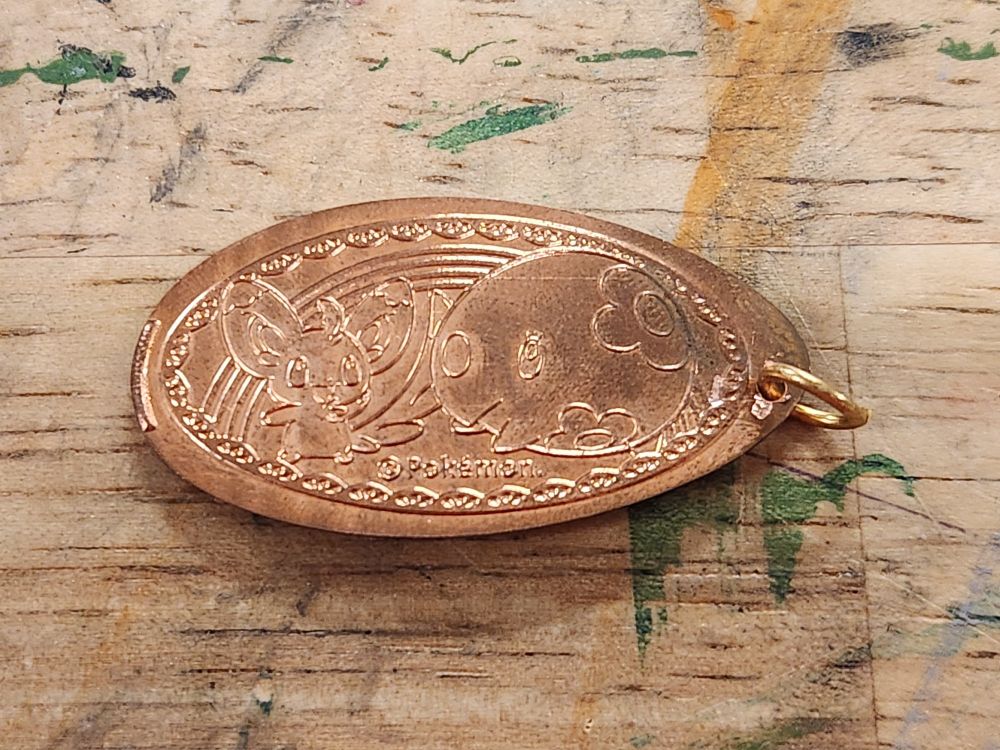 A pressed copper coin with Minccino and munna from pokemon indented on it. It has a rainbow indent in the background and a lacy indent border around it. I drilled a hole in a part on the right edge without any design where the penny stretched and attached a jump ring.