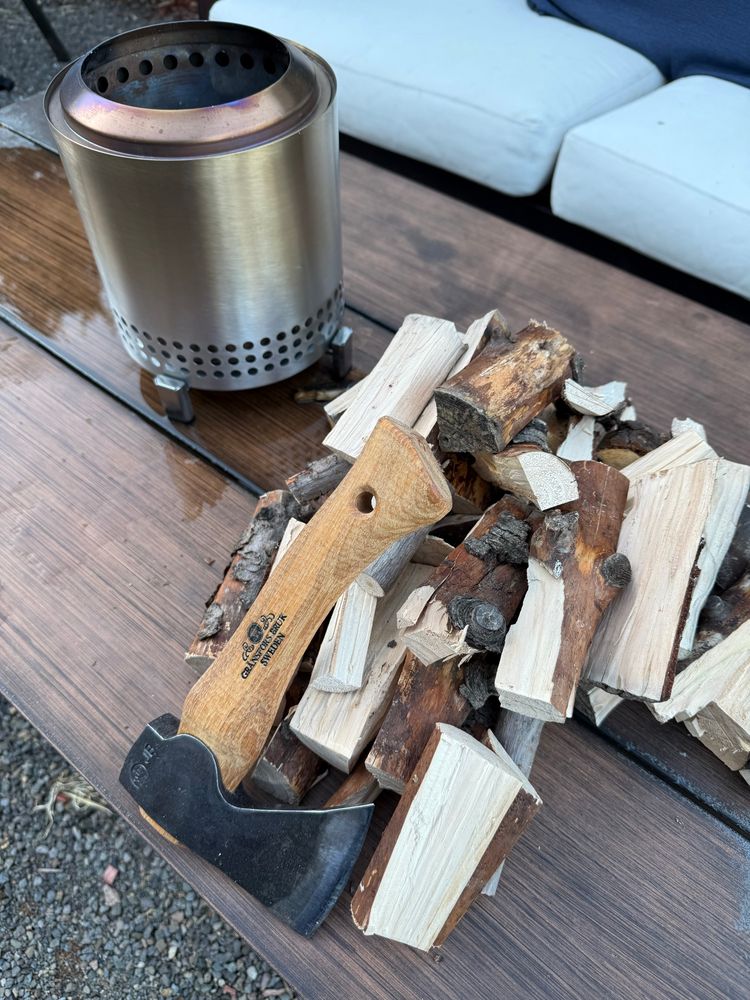 Shows a hand axe resting on wood near a fire pit. 