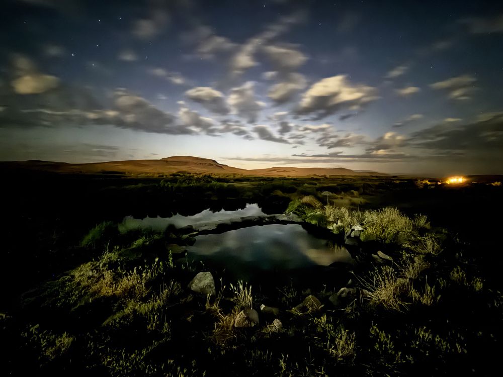 Shows Willow Creek Hot Springs in the foreground with hills behind lit up by moonlight and lights from a camp nearby. There are wispy clouds in the sky and vegetation around the spring. 