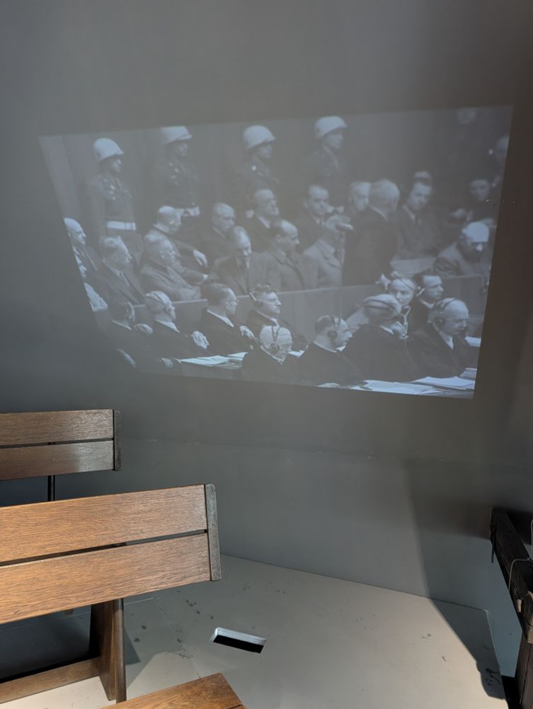Photo des bancs exposés dans le mémorial des procès de Nuremberg, avec une image des procès en fond.