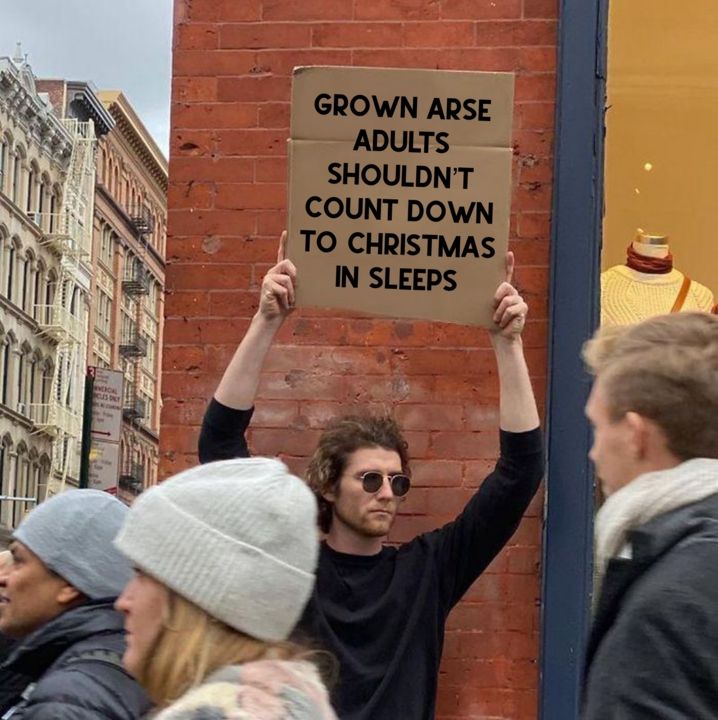A man holds up a cardboard sign on a street corner. It says 'Grown arse adults shouldn't count down to Christmas in sleeps'.