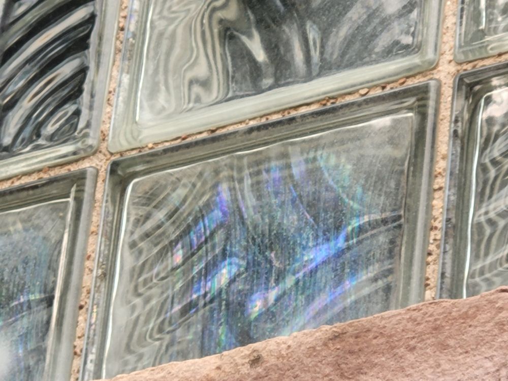 This picture shows a portion of a glass block window. One block is iridescent with blues, greens, and purple shining through. The other blocks have light and dark areas swirling through the glass. The window sill and the grout between the blocks is rough-looking and light terra cotta in color.