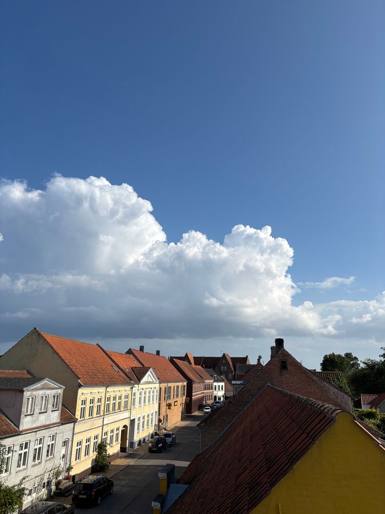 This is a lovely view of what appears to be a traditional Danish town centre, with characteristic colourful buildings featuring red clay tile roofs. The architecture shows the classic Scandinavian style with multi-storey buildings in yellow, white, and other pastel colours lining a quiet street. The dramatic sky filled with puffy white cumulus clouds against the deep blue backdrop creates a beautiful contrast with the historic buildings below. The perspective suggests this photo was taken from an elevated position, perhaps from an upper floor window or rooftop, looking down at the charming streetscape with its mix of residential and possibly commercial buildings typical of Danish market towns.​​​​​​​​​​​​​​​​
