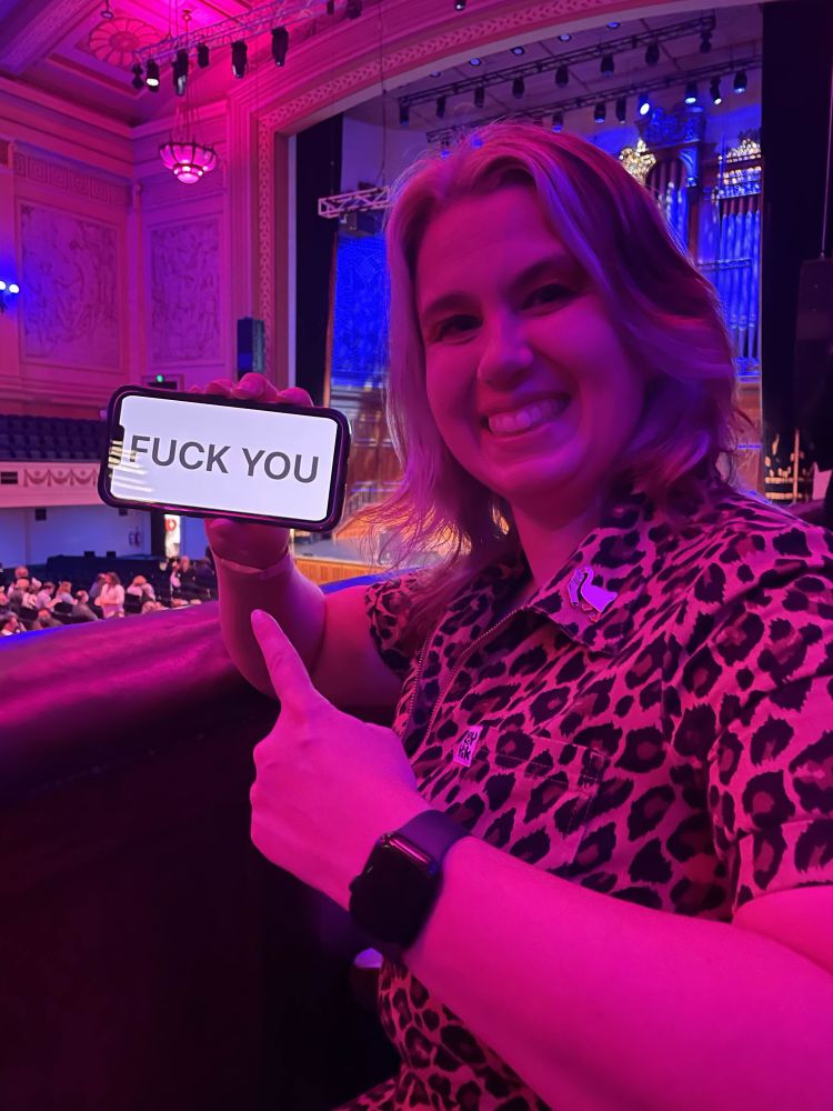 A cheeky 5’2 woman in a leopard print boiler suit pointing to her phone that just says ‘fuck you’