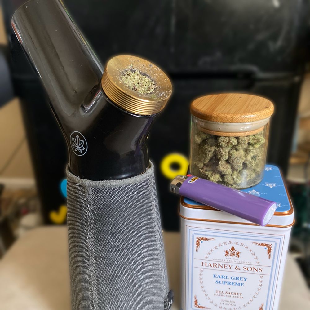 A photo of a ceramic glass and brass water bong wrapped in a grey cozy cover. On the right is a white tea tin with light blue and copper accents that reads Harney and Sons Earl Grey Supreme Tea Sachet. On top of this tea tin is a purple bic lighter. Next to the lighter is a bamboo and glass jar with weed inside of it. 