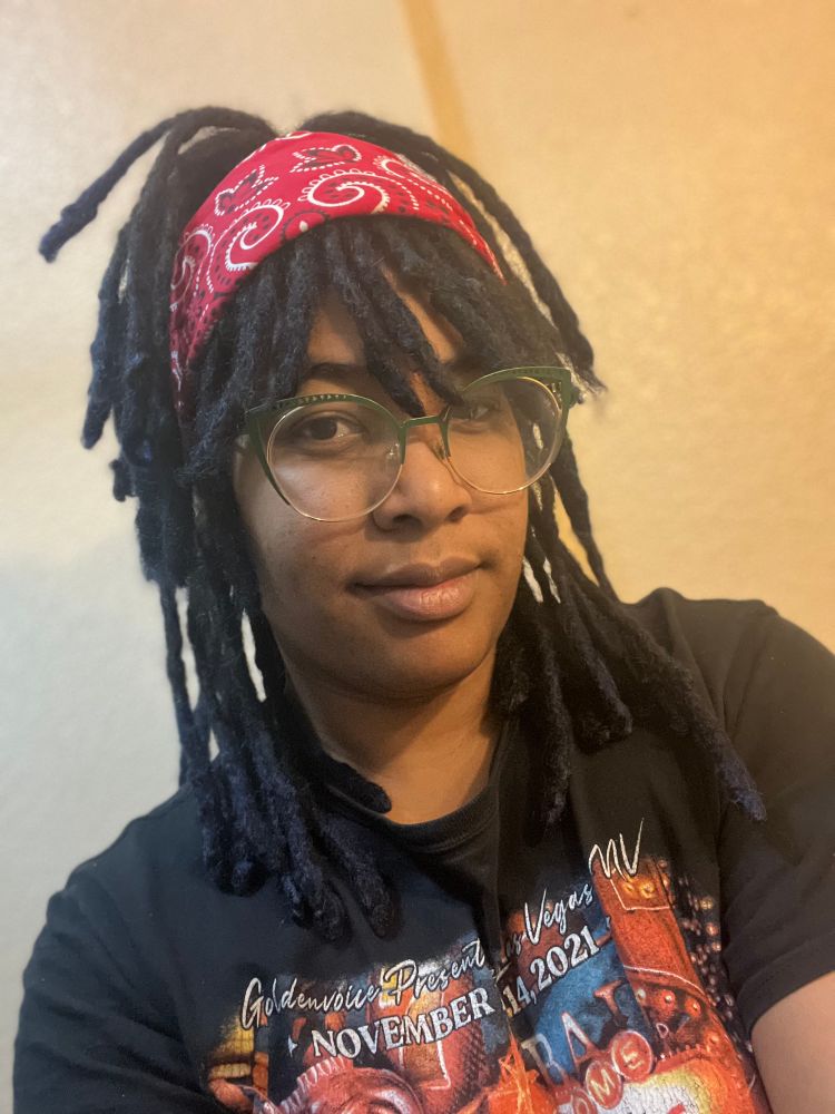 A black woman poses for the selfie. She is standing against a beige to cream ombré wall. She is wearing a black shirt with a red orange white and blue graphic on the front that is mostly cut off in the photo with the date November 14,2021 visible. She has collarbone length locs that are dyed different shades of dark blue. She has on a red paisley patterned bandana, tying up half her locs in a high ponytail, and her bangs are gently resting on her gold and green framed cat eye glasses. 