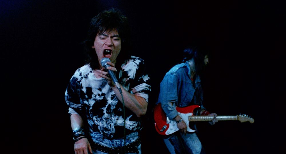 Diamond Yukai as Hiro from Tokyo Pop (1988). He's dressed as a rocker and singing, screaming at a microphone. There's a guitar player behind him, playing a red guitar.