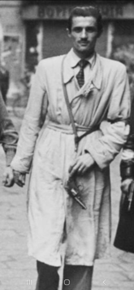 A zoomed in portion of the above photo of Italian partisans.

A tall man with slicked hair and a moustache, wearing a trenchcoat, with a strap crossing his chest and a handgun apparently hanging from the strap. His hand is lightly holding the gun, presumably to stop it swinging.

At first glance, however, it looks like he is holding the gun with 3 fingers by the handle with the barrel hanging down.