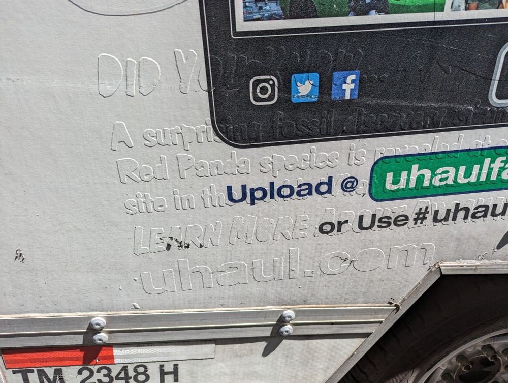 Photo of the side of a u haul truck. You can just make out raised text with fun facts about red pandas but it's covered in white and not very legible, from another (inferior) design on top 