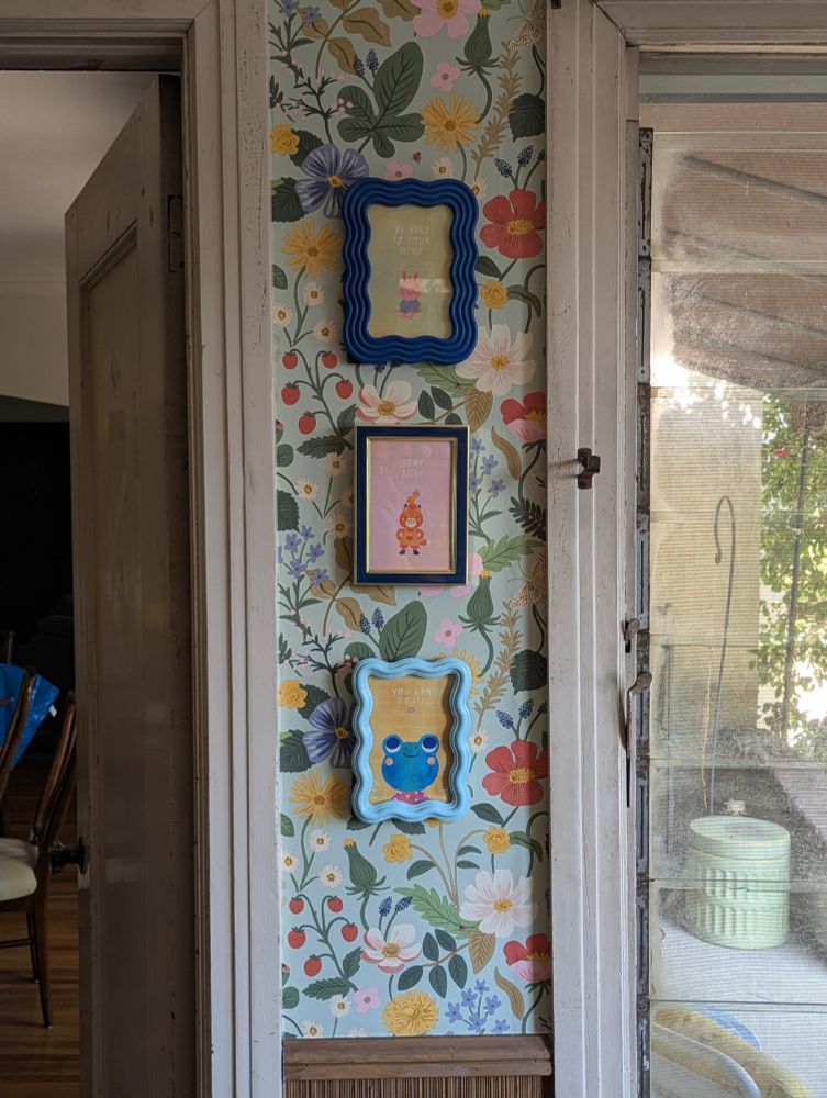 Photo of a section of wall between a window and a door. There are three frames prints hung on the wall.