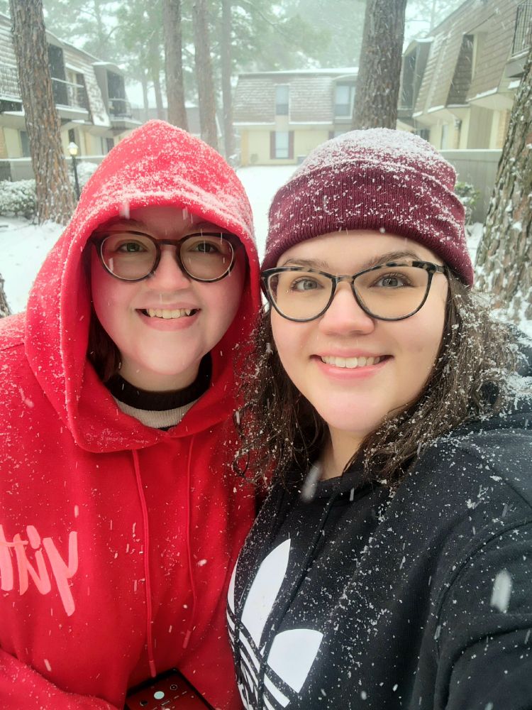 two brunette, four-eyed, green-eyed, big-grinned sisters standing in the snow, one with a red yitty hoodie and the other in a black adidas hoodie and beanie.