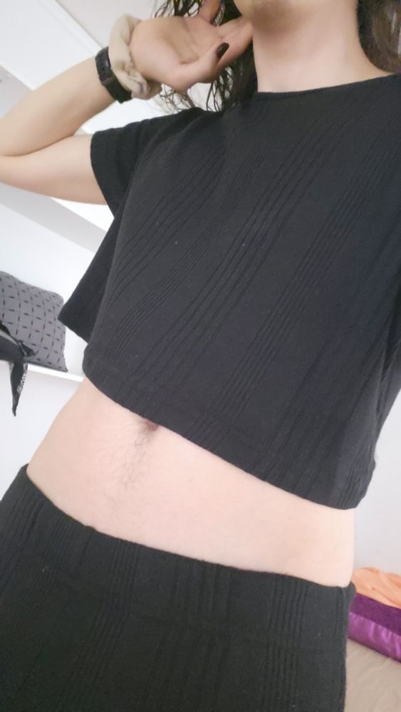 Picture of me, a latino enby, from my neck to my hips. I'm wearing a matching black crop top and pajama pant set. I'm raising my left arm to my neck, raising the crop top to expose the tops of my hips, waist, and navel area
