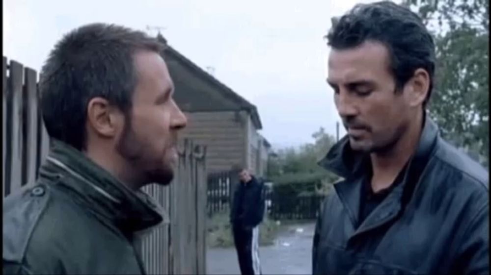 two men are standing next to each other in front of a wooden fence .