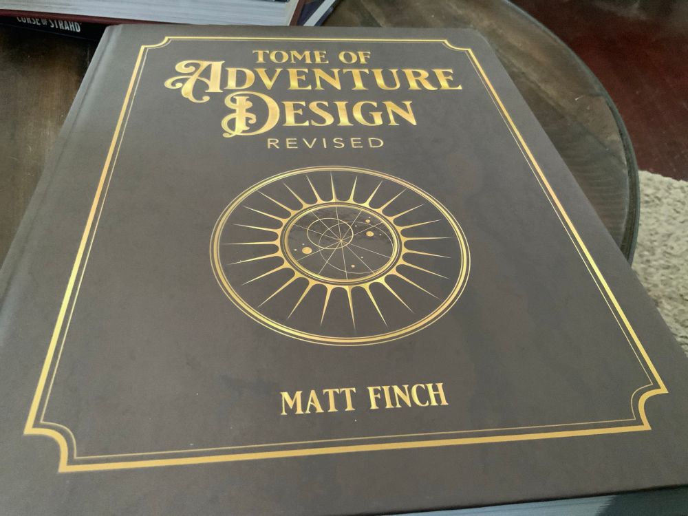 A large hardback book titled "Tome of Adventure Design Revisited" by Matt Finch. It has a simply designed cover with faux gold trim and a circular design element in the center. 