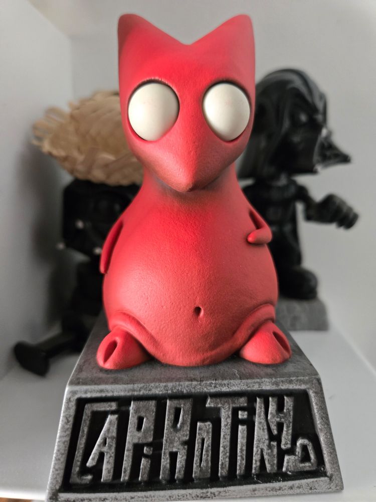 Uma estatueta de um personagem estilizado e minimalista, com corpo vermelho, olhos grandes e brancos e uma pose descontraída. A figura está em cima de uma base de metal que tem o texto "Capirotinho" gravado.