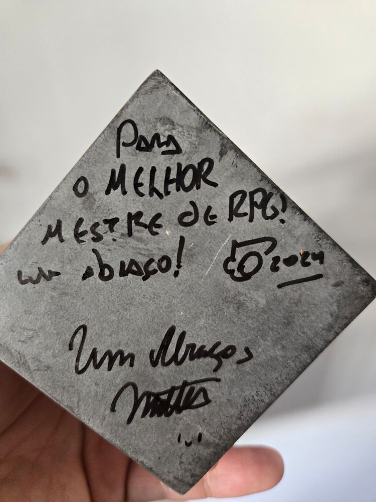 Um objeto de metal com uma dedicatória escrita à mão em português, que diz: "Para o MELHOR MESTRE DE RPG! Um abraço! 2024". Logo abaixo, a mensagem é assinada: "Um abraço, Mattos :)". O fundo é neutro, e a peça está sendo segurada por uma mão. A escrita é feita com uma caneta preta e exala carinho e gratidão.
