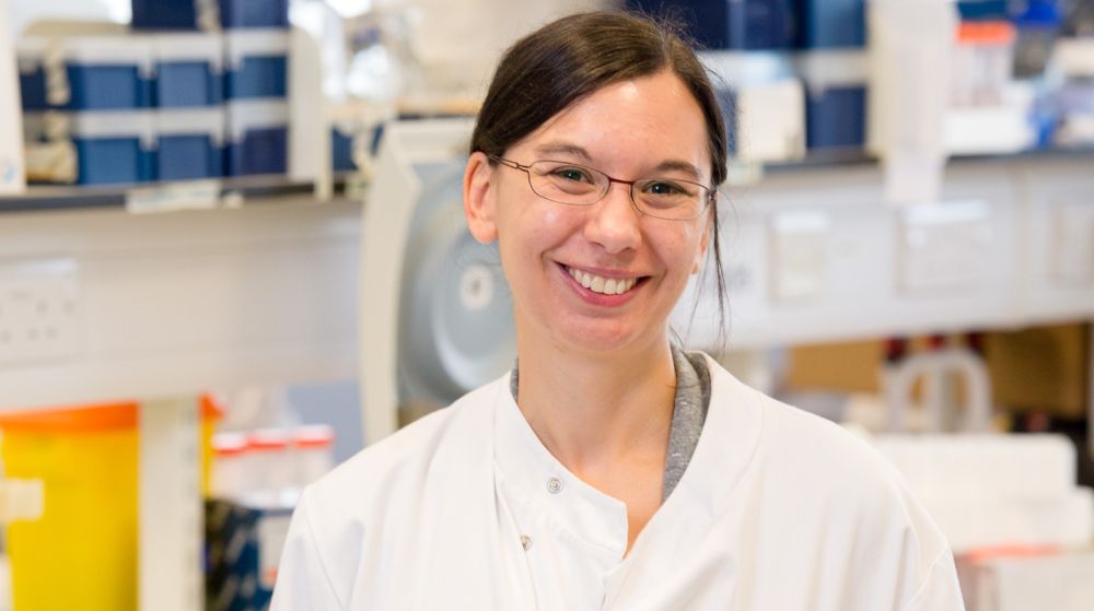 A head and shoulders portrait photograph of a smiling Dr Megan Macleod stood in the lab wearing a white lab coat.