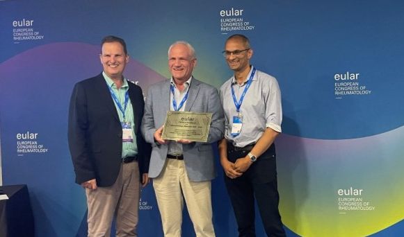 Professor Iain McInnes (Head of College of MVLS), Ronald Van Vollenhoven (Research Chair of EULAR), Professor Neil Basu (Sii Professor of Musculoskeletal Medicine & Vasculitis) stood in front of a pull-up banner with EULAR branding, with Ronald holding the presentation plaque.
