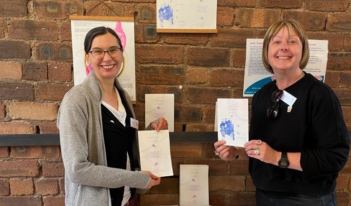Dr Megan Macleod and Jane Topping stood in front of an indoor feature wall made of brick while holding up the art produced. The art also decorates the wall behind them.