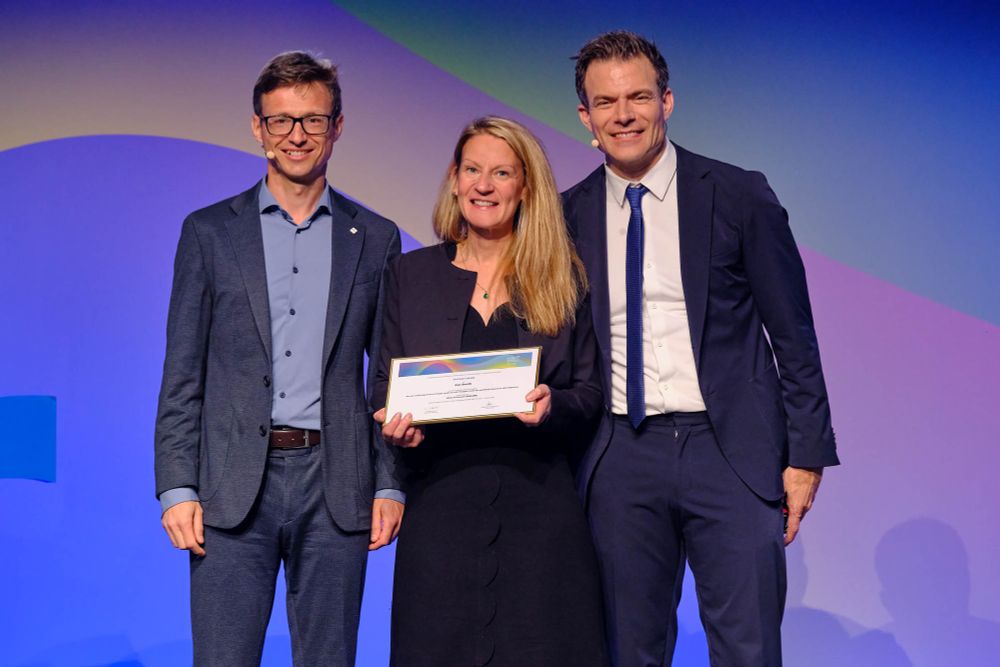 Dr Eve Smith stood on the stage at the EULAR Conference holding a certificate of her award, with those presenting the award stood on either side.