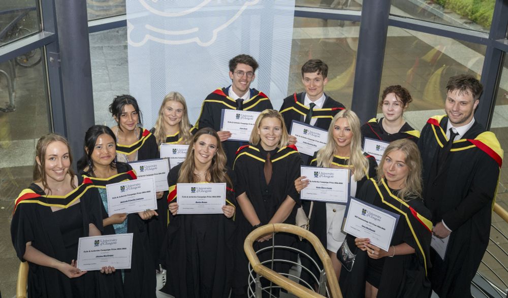 Immunology graduates stood together holding prize winner certificates in the Sir Graeme Davies stairwell in front of the large windows showing outside behind them.
