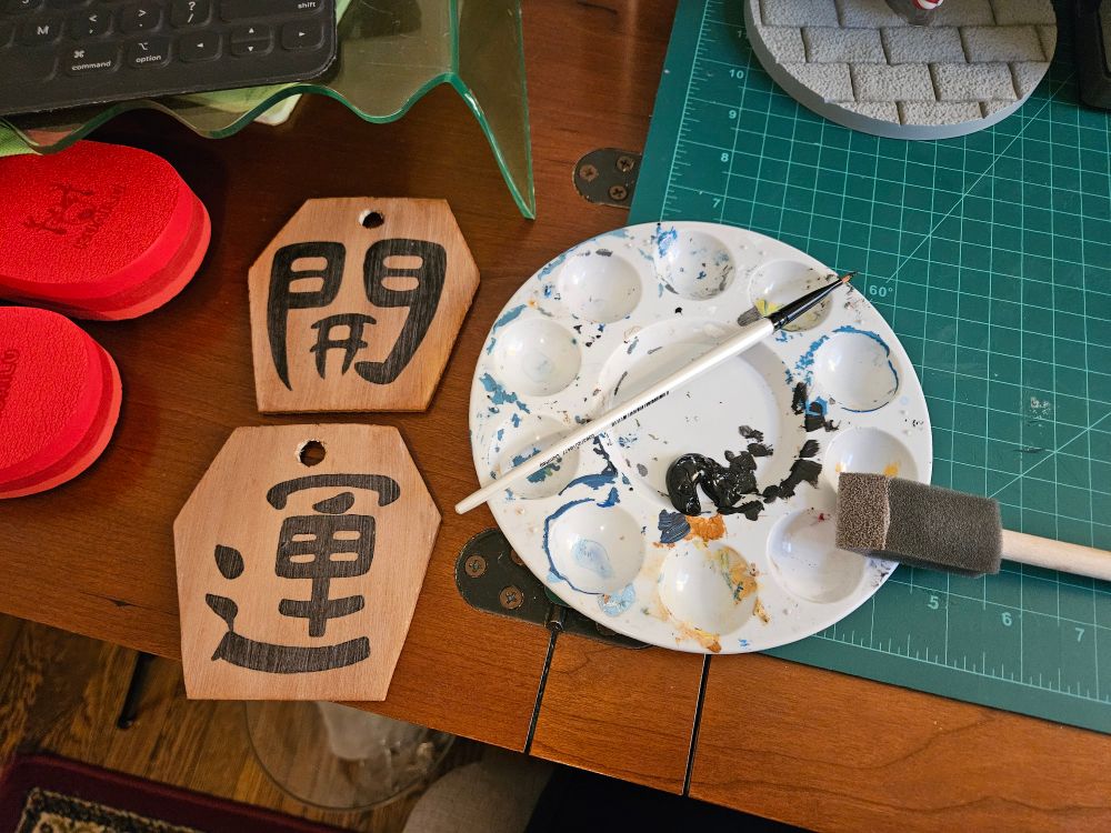 Two omamori with kanji sit next to a paint pallette and brushes