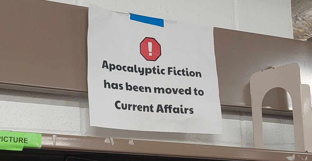 A sign on a metal bookshelf that says "Apocalyptic Fiction has been moved to Current Affairs"