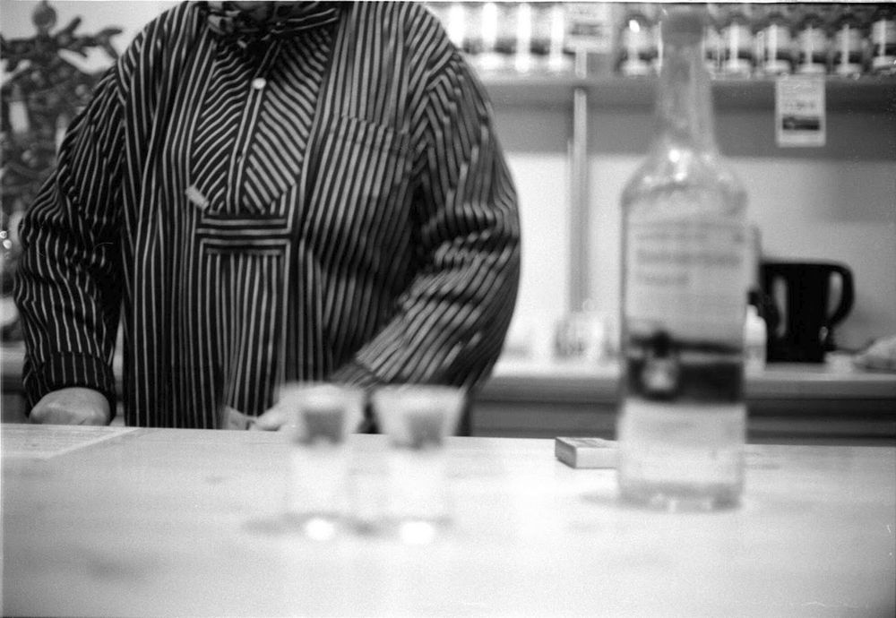 Black and white image of a guy at a schnaps place, not focusing on the schnaps because why would I do that, I want to focus on the half cut-off dude in the background instead. Yes, that's what I wanted. 
Agfa APX100 on a Revue 400SE
