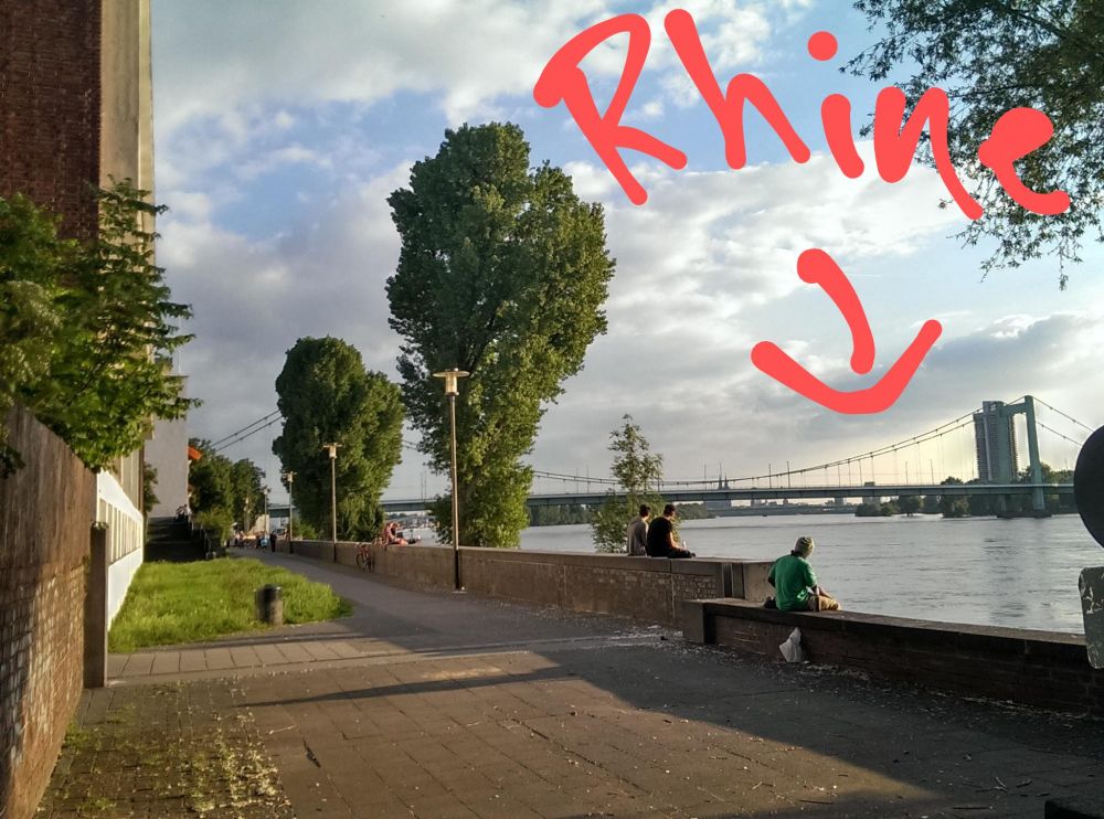 View from the North of Cologne near Stammheim and towards the Mülheim bridge, with a big "Rhine" pointing at the Rhine, Cologne cathedral far off in the distance