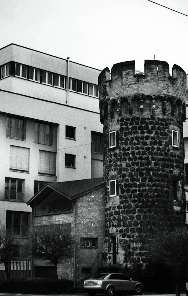 Black and white photo from a medieval tower next to two modern buildings in Cologne, Germany. Car parked out front, taken on a Minolta X-500 film SLR on Agfa APX 100, unknown lens.