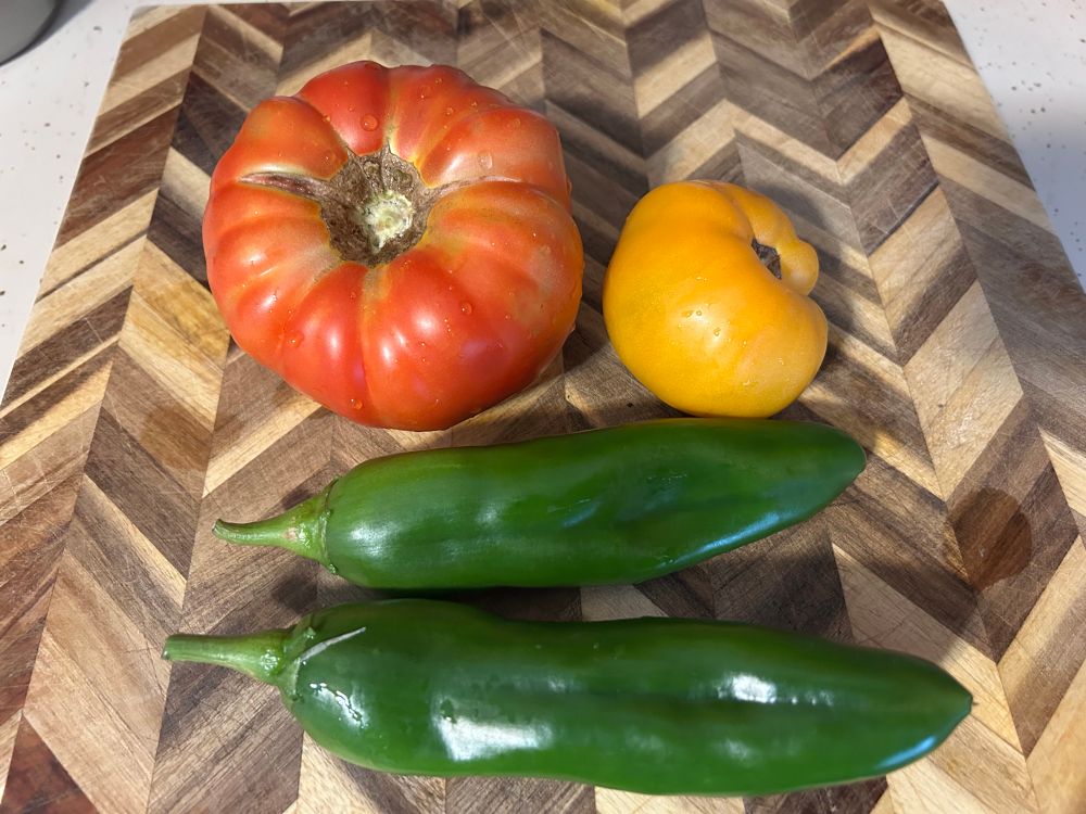 A large beefsteak tomato, a smaller Wyche’s Yellow tomato and two Anaheim peppers on a wooden herringbone cutting board 