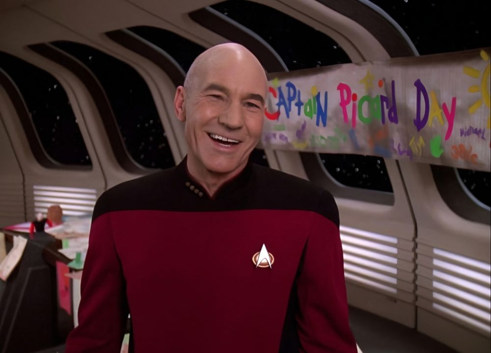 The image depicts Captain Jean-Luc Picard standing in a futuristic spaceship, the Federation starship USS Enterprise (NCC-1701-D). He is wearing a red and black Starfleet uniform with the distinctive insignia on the left side of his chest. This is the standard command uniform from "Star Trek: The Next Generation," featuring the captain's rank insignia of four gold pips on the collar. Behind him, a banner with colorful text reads, "Captain Picard Day," celebrating one of the greatest captains in the galaxy. The background features large windows with a view of space, and other crew members and equipment are visible. Captain Picard is smiling, and the overall atmosphere is cheerful and festive.