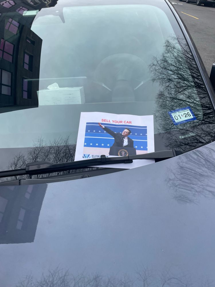 Picture of a cars windshield with a flyer under the wiper blade with a picture of Elon Musk doing the Nazi salute and the words “Sell your Car”

