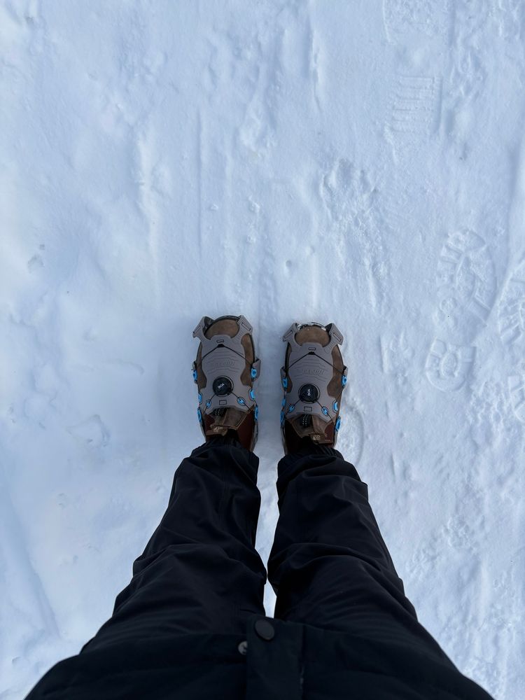 A downward view of my legs in black hiking pants, grey ice cleats attached to my boots, on a snow-covered trail. 