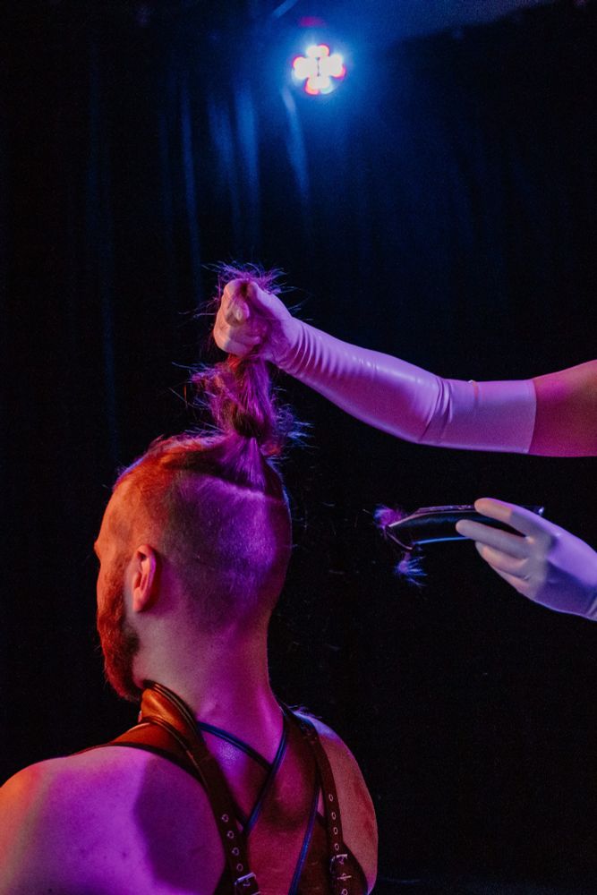 Cymbelin in a leather corset is kneeling in front of their wife and owner with their back towards the camera. Their owner holds clippers while wearing shiny long white Latex gloves and is about to cut off Cymbelin's long hair
